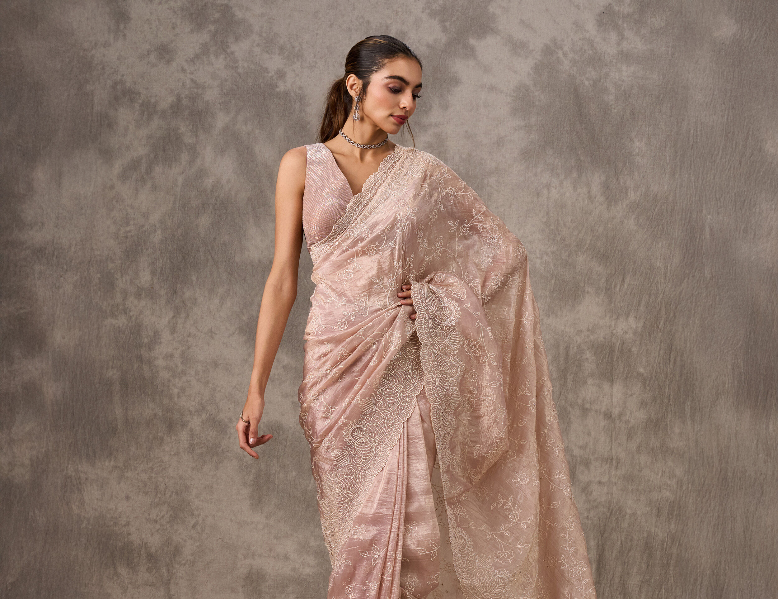 Woman modeling a sheer, intricately embroidered nude pink saree with a matching sleeveless blouse for elegant styling.